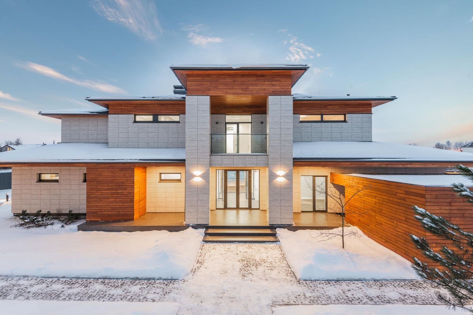 Warmly lit two-story modern home with wood and stone siding on a snowy winter evening.